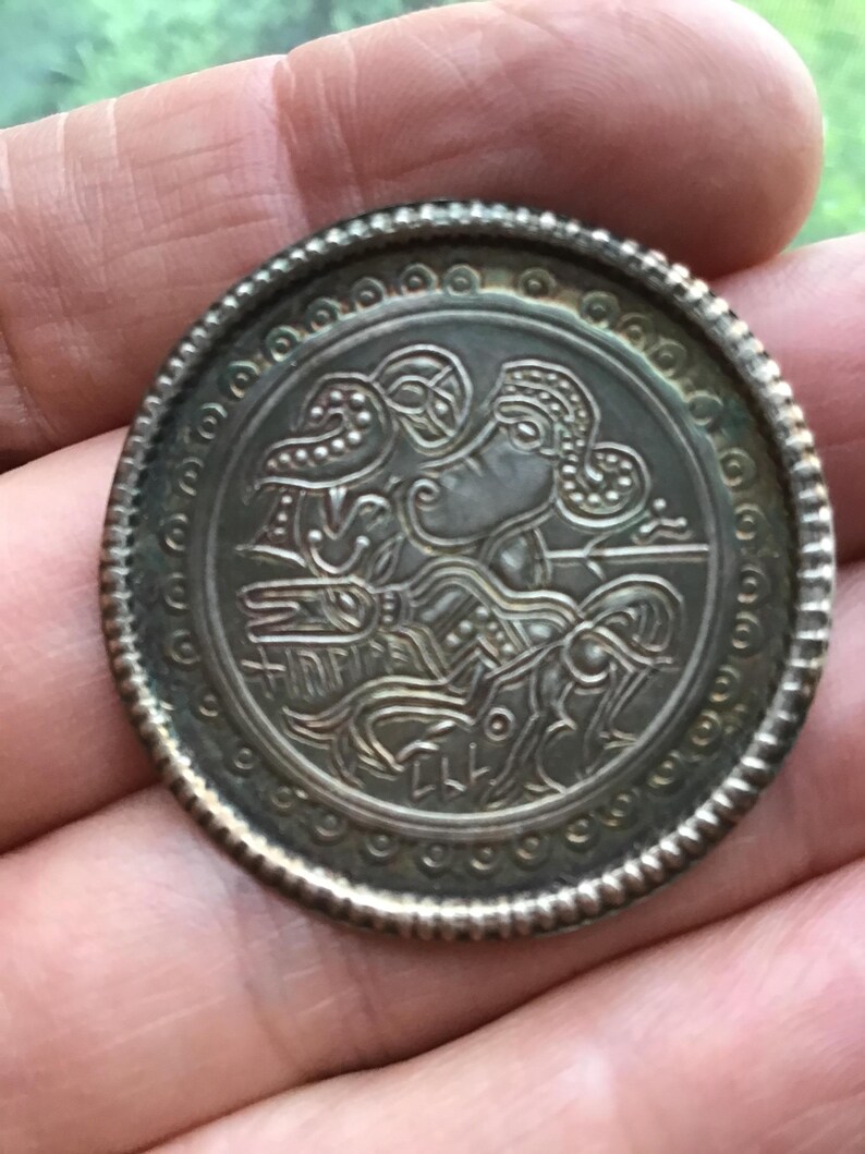 May include: A silver-colored, round coin with an intricate embossed design. The central image features a stylized figure with detailed patterns, surrounded by a decorative border. The coin's edge has a textured, beaded pattern.