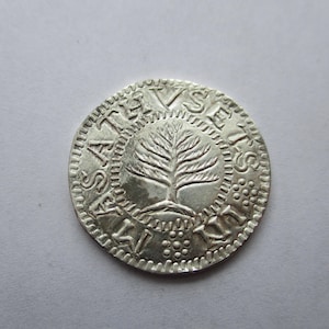 May include: A silver coin with a detailed tree design at the center, surrounded by an inscription. The coin's edge features a textured pattern. The coin appears to be an antique or historical artifact.