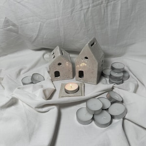 Op de afbeelding: Twee witte keramische huisjes met een gespikkelde afwerking, elk met een klein gaatje voor een theelichtje. Een brandend theelichtje bevindt zich in een van de huisjes. Er zijn meerdere witte theelichtjes op een wit oppervlak.