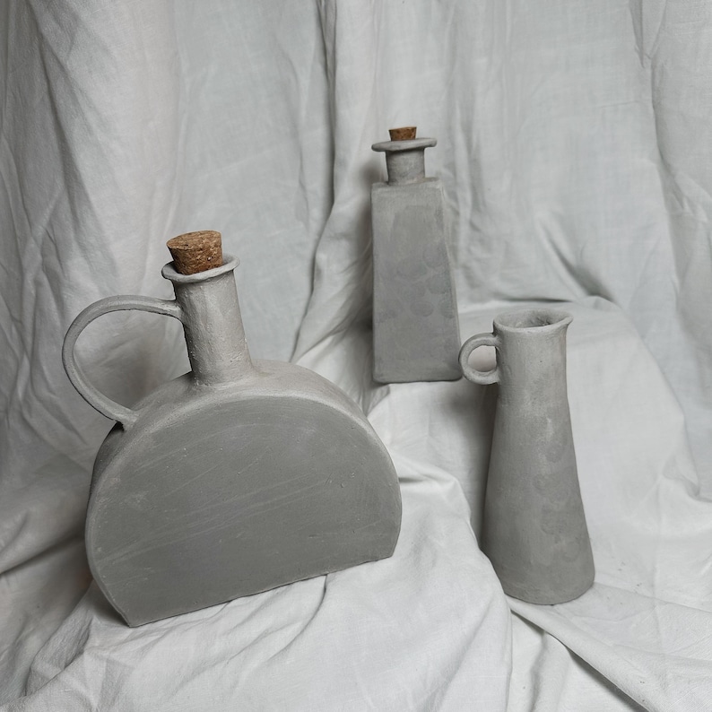 May include: Three gray ceramic vases with cork stoppers. The vases are of different shapes and sizes. The largest vase has a rounded body and a handle. The middle vase is rectangular. The smallest vase is tall and slender.
