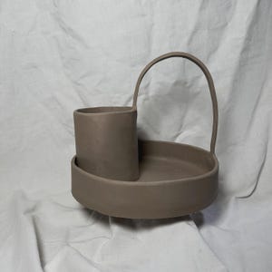 Puede incluir: Una bandeja de cerámica marrón con asa y un recipiente cilíndrico separado. La bandeja y el recipiente son ambos sin esmaltar.