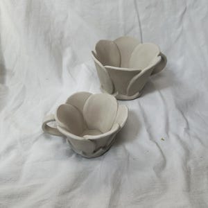 May include: Two ceramic flower-shaped cups, one larger than the other, both in a light beige color. The cups are unglazed and have a simple, rustic design.