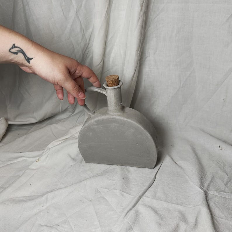 May include: A gray ceramic bottle with a cork stopper and a handle. The bottle is sitting on a white fabric surface.