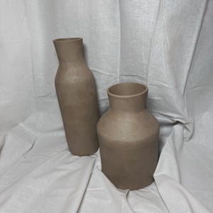 以下が含まれることがあります： 異なるサイズと形の2つの茶色の陶器の花瓶。大きな花瓶は背が高く、細身です。一方、小さな花瓶は背が低く、幅広です。