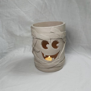 Puede incluir: Un portavelas de Halloween de cerámica beige con forma de momia. La momia tiene ojos recortados y una boca sonriente, con vendas alrededor del cuerpo. Una vela encendida está dentro, emitiendo un brillo cálido.