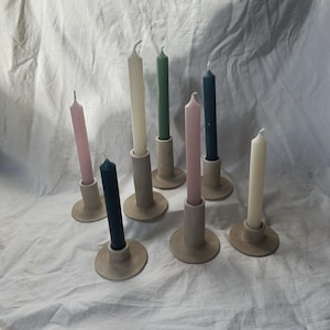 Puede incluir: Colección de siete velas cónicas de varios colores, incluyendo rosa, verde y azul marino, cada una colocada en un portavelas cilíndrico de tono neutro. Las velas están dispuestas sobre una superficie blanca texturizada, creando una estética minimalista.