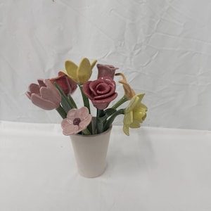 Op de afbeelding: Een keramisch bloemstuk met tulpen, rozen en narcissen in roze, rood en geel. De bloemen staan in een kleine, gebroken witte vaas, wat een decoratieve bloemenpresentatie creëert.