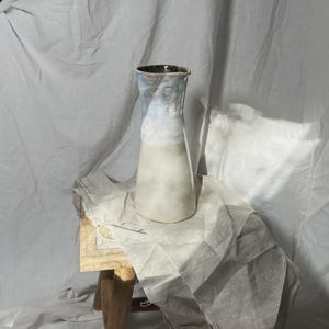 Può includere: Un vaso di ceramica bianca con una glassa blu chiaro e un bordo marrone. Il vaso è appoggiato su un panno di lino bianco su uno sgabello di legno.