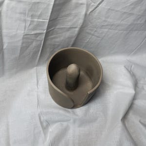 K&ouml;nnte beinhalten: Ein handgefertigter, grauer Keramik-Ringhalter mit einem zylindrischen Mittelpfosten und einem geschwungenen, offenen Design. Der Halter ist vor einem wei&szlig;en, strukturierten Hintergrund platziert und zeigt seine minimalistische &Auml;sthetik.
