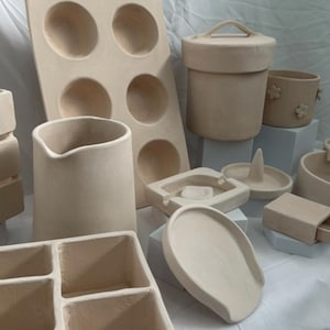 May include: A collection of handmade, beige ceramic items. Includes a pitcher, a container with a lid, a tray, a divided tray, a ring holder, a small ashtray, and a mold with six circular indentations. The items are arranged on white and light blue blocks.