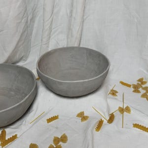 Puede incluir: Dos cuencos de cerámica gris sobre una superficie de tela blanca con pasta de lazo esparcida.