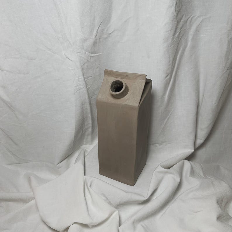 May include: A light brown ceramic vase shaped like a milk carton.
