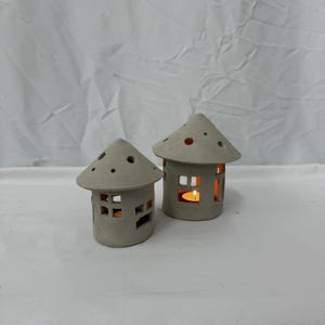 May include: Two small, light grey ceramic house-shaped candle holders. Each has a conical roof with small holes and rectangular windows. One candle holder has a lit candle inside, casting a warm glow.