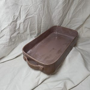Op de afbeelding: Een rechthoekige keramische ovenschaal in een gedempte mauve kleur. De schaal heeft afgeronde hoeken, een gladde binnenkant en twee gebogen handvatten aan de zijkanten. Het ontwerp is eenvoudig en functioneel, geschikt om in te bakken en te serveren.