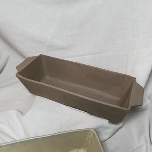 Może przedstawiać: Prostokątna, jasnobrązowa ceramiczna donica o lekko trapezowym kształcie. Posiada dwa małe uchwyty po bokach. Donica przeznaczona jest do sadzenia kwiatów lub ziół.