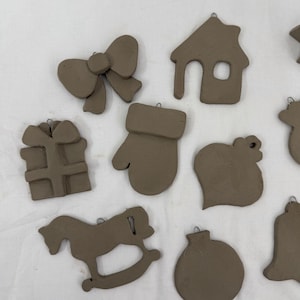 May include: A collection of unglazed clay Christmas ornaments in various shapes, including a bow, house, gift, mitten, heart, rocking horse, and bell. Each ornament has a small loop for hanging. The ornaments are a neutral brown color.