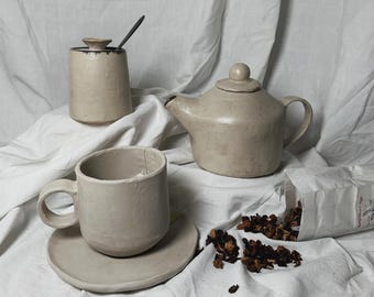 May include: A beige ceramic teapot, sugar bowl, and teacup with saucer. The teapot has a rounded body and a small spout. The sugar bowl has a lid. The teacup has a handle and a saucer. There are loose tea leaves scattered on a white fabric surface.