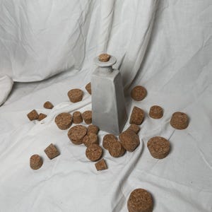May include: A gray ceramic bottle with a cork stopper surrounded by a scattering of cork stoppers on a white fabric surface.