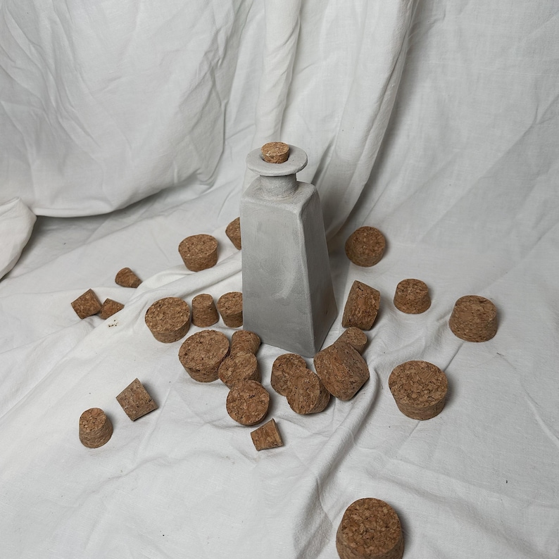 May include: A gray ceramic bottle with a cork stopper surrounded by a scattering of cork stoppers on a white fabric surface.