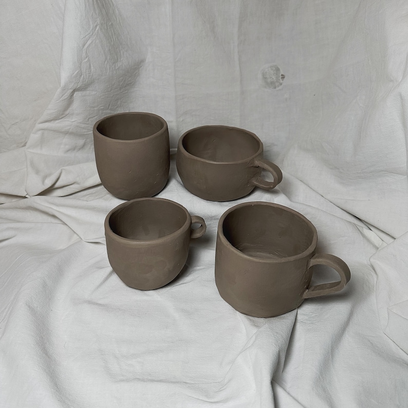 May include: Four brown ceramic mugs of varying sizes, two with handles, on a white fabric background.