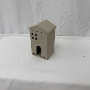 Puede incluir: Un pequeño recipiente de cerámica gris con forma de casa y techo extraíble. La casa tiene un cuerpo rectangular con una puerta arqueada y cuatro ventanas cuadradas. El techo es inclinado y el diseño es minimalista.