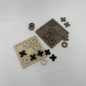 Könnte beinhalten: Zwei Tic-Tac-Toe-Spielsets. Ein Set ist beige mit schwarzen X's und weißen O's. Das andere Set ist braun mit braunen X's und O's. Zusätzliche X's, O's und ein Satz von acht Ringen sind um die Spielbretter verstreut.