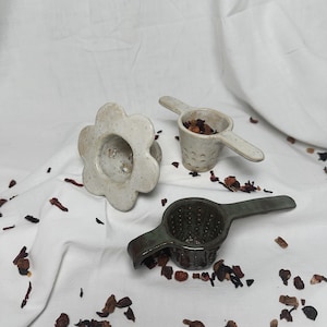 Puede incluir: Tres infusores de té de cerámica. Uno es un infusor de forma de flor blanca, otro es un infusor cónico blanco con asa, y el tercero es un infusor cónico verde oscuro con asa.