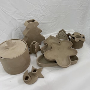 Peut inclure: Ensemble d'objets en céramique non émaillée, comprenant un pot avec couvercle, un vase en forme de sapin de Noël, un plateau en forme de bonhomme en pain d'épices et d'autres formes festives. Les articles sont disposés sur une surface blanche.