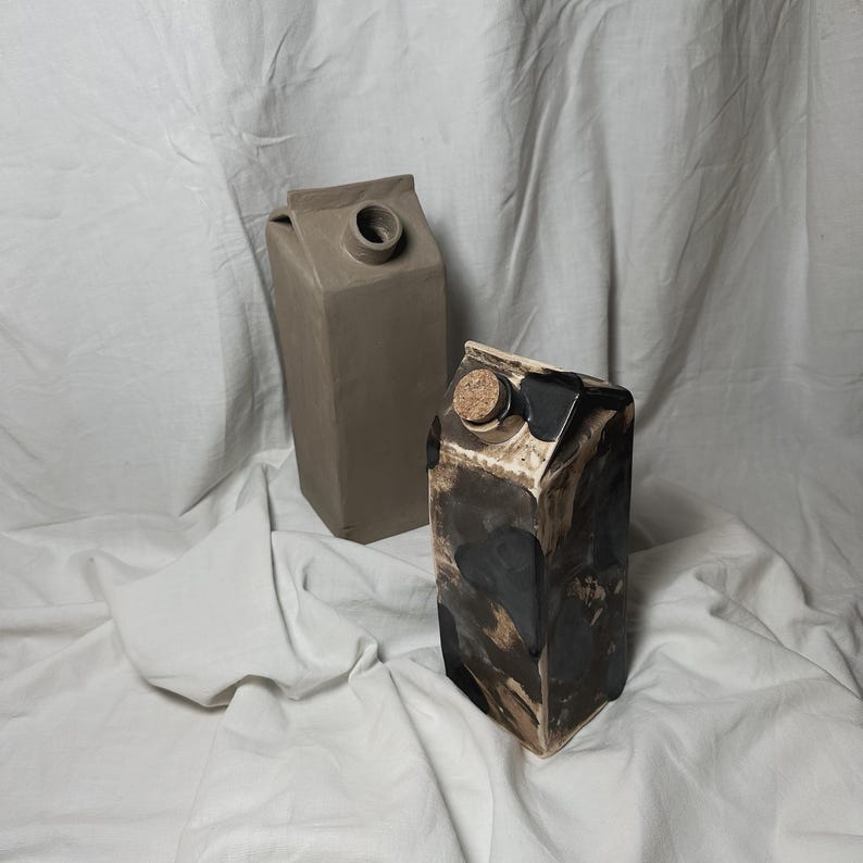 May include: Two ceramic milk carton vases, one is brown and the other is black with white and brown splatter paint.