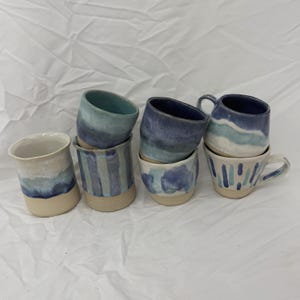 Può includere: Una collezione di tazze e tazze in ceramica fatte a mano in varie tonalità di blu e bianco. La ceramica presenta disegni unici, tra cui strisce e motivi ad acquerello. Le tazze hanno un'estetica rustica e artigianale.