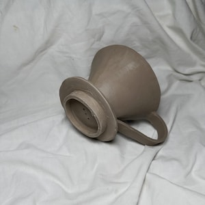 May include: A brown ceramic coffee filter with a handle and a round base with three holes. The filter is designed for pour-over coffee brewing.