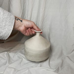 Puede incluir: Un jarrón de cerámica blanco con cuerpo redondeado y parte superior ancha y plana. El jarrón tiene un glaseado sutil que crea un efecto de capas.