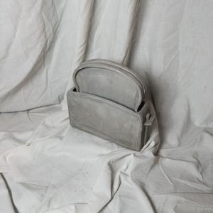 Peut inclure: Un sac à main en céramique gris au design unique. Le sac a un dessus à charnière et une poignée.