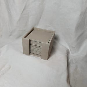 May include: A set of square, light beige ceramic coasters in a matching holder. The coasters are stacked neatly inside the holder, which has a simple, minimalist design. The set is designed to protect surfaces from drink condensation.