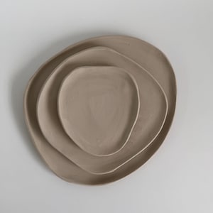 May include: A set of three nested plates in a matte beige finish, each featuring an irregular, organic, and asymmetrical shape with smooth, slightly raised edges. The plates are stacked centrally, displaying a consistent earthy tone throughout.