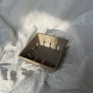 May include: A square, beige ceramic basket with vertical slots along the sides. The basket is set against a white, textured background. The basket's design suggests it could be used for storage or display.