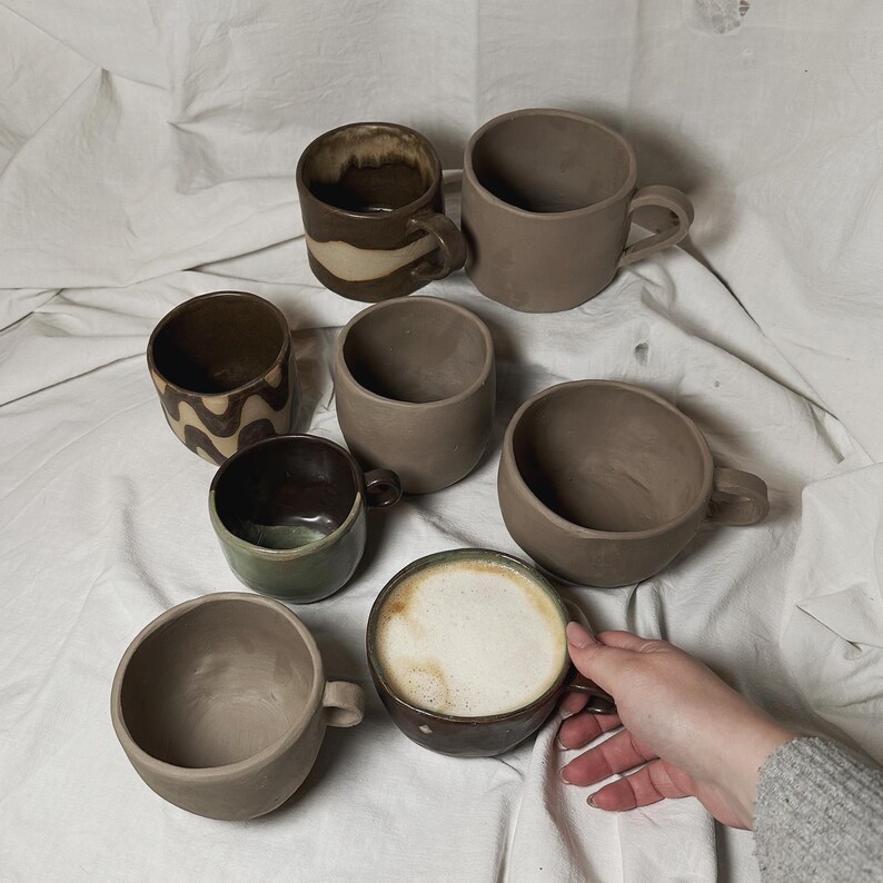 May include: A collection of seven ceramic mugs in various shades of brown and beige. Some mugs have a textured surface, while others have a smooth finish. One mug is filled with a light brown liquid, possibly coffee or tea.