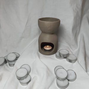 May include: A brown ceramic oil burner with a small tea light candle burning inside. The burner is surrounded by a pile of white tea light candles.