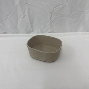 May include: A small, handmade, light grey ceramic bowl with rounded corners. The bowl has a simple, minimalist design and is likely used for serving or storage. The neutral colour makes it versatile for various home decor styles.
