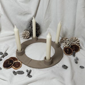 May include: A brown ceramic candle holder in the shape of a ring with four candle slots. The holder is decorated with a rustic, textured finish. Four white candles are placed in the slots.