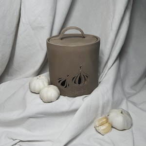 May include: A brown ceramic garlic keeper with a lid and a handle. The keeper has two cutouts in the shape of garlic cloves. The word "Garlic" is written on the front of the keeper. There are three whole garlic bulbs and two cloves on a white fabric surface.