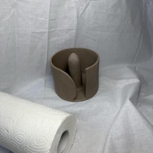 K&ouml;nnte beinhalten: Ein brauner Papiertuchhalter aus Keramik mit zentraler vertikaler St&uuml;tze. Eine Rolle wei&szlig;er Papiert&uuml;cher mit sich wiederholendem Muster befindet sich im Vordergrund. Der Halter hat ein geschwungenes Design und ist vor wei&szlig;em Hintergrund platziert.