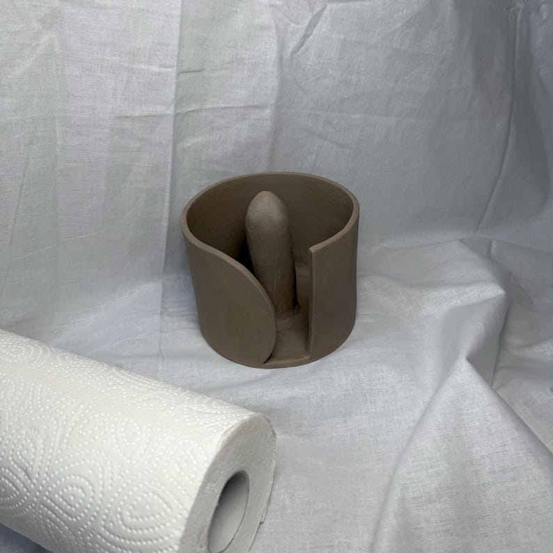 K&ouml;nnte beinhalten: Ein brauner Papiertuchhalter aus Keramik mit zentraler vertikaler St&uuml;tze. Eine Rolle wei&szlig;er Papiert&uuml;cher mit sich wiederholendem Muster befindet sich im Vordergrund. Der Halter hat ein geschwungenes Design und ist vor wei&szlig;em Hintergrund platziert.
