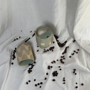 Könnte beinhalten: Zwei Keramikbecher mit einzigartigen Designs. Einer ist ein hellgrüner Zylinder mit einem Gittermuster, der andere hat ein hellblaues und beiges Wellenmuster. Kaffeebohnen sind um die Becher auf einem weißen Tuch verstreut.
