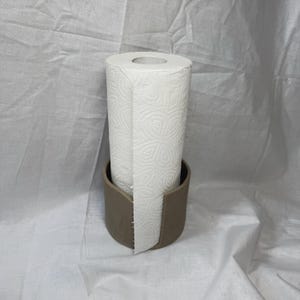 K&ouml;nnte beinhalten: Eine wei&szlig;e K&uuml;chenpapierrolle mit dekorativem Pr&auml;gemuster in einem taupefarbenen Keramikhalter. Der Halter hat ein gebogenes Design f&uuml;r einfachen Zugriff. Der Hintergrund ist ein wei&szlig;er Stoff. Der Halter ist etwa 15 cm hoch.