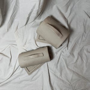 May include: Two beige ceramic mugs with rectangular bases and attached handles. The mugs are placed on a white, wrinkled fabric background. The mugs have a minimalist design.