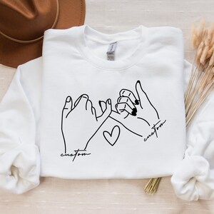 Puede incluir: Sudadera blanca con un dibujo de l&iacute;nea negra de dos manos entrelazadas con un coraz&oacute;n en el medio. El texto "custom" est&aacute; escrito debajo de cada mano.
