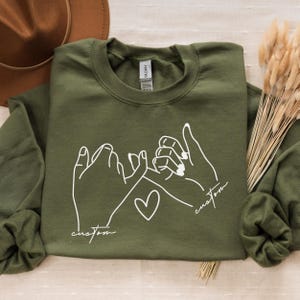 Puede incluir: Una sudadera verde con un contorno blanco de dos manos haciendo una promesa me&ntilde;ique. El texto "custom" est&aacute; escrito debajo de cada mano.