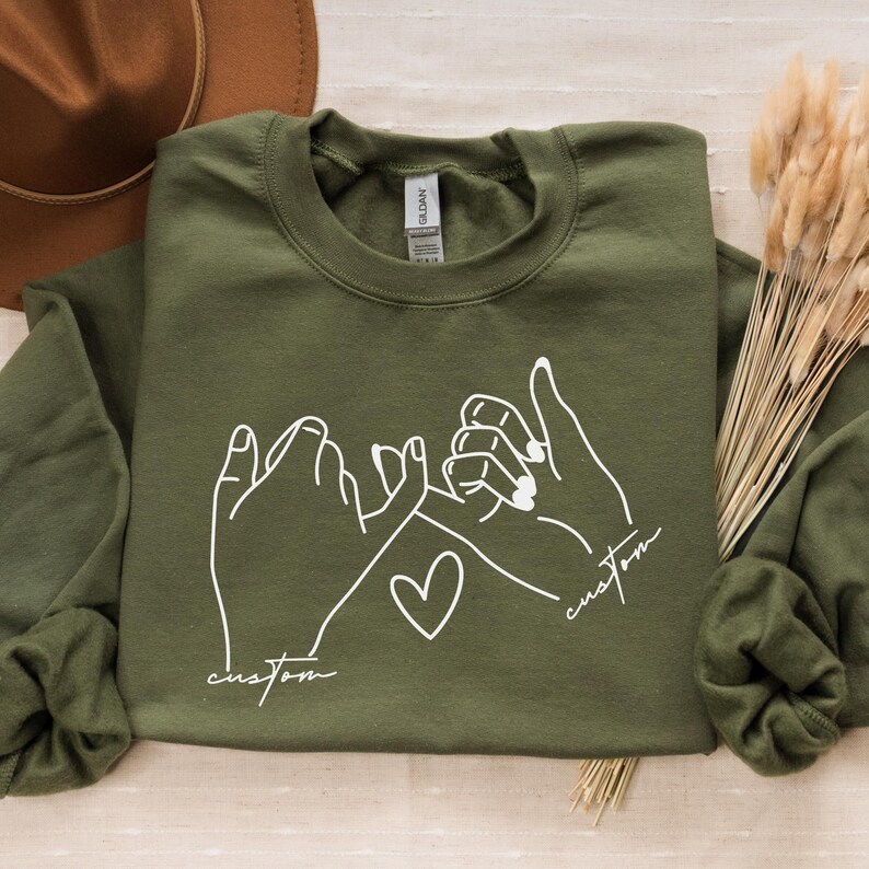Puede incluir: Una sudadera verde con un contorno blanco de dos manos haciendo una promesa me&ntilde;ique. El texto "custom" est&aacute; escrito debajo de cada mano.