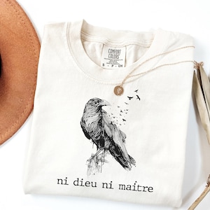 May include: Cream-colored t-shirt featuring a detailed black raven illustration with the phrase "ni dieu ni ma&icirc;tre" printed below. A gold necklace and a brown hat are also visible. The shirt is made of Comfort Colors material.
